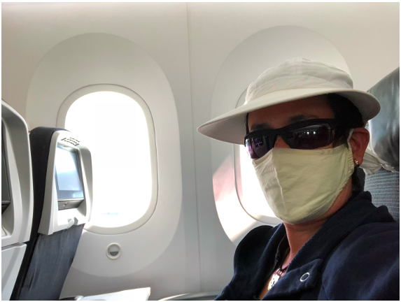 Travel during COVID: ILONA POSNER seated in an airplane wearing a face mask, sun glasses, and a white hat.